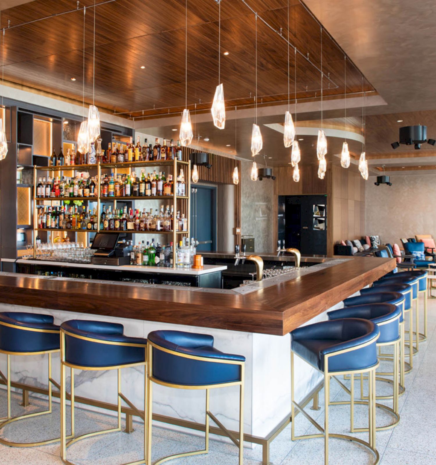 Modern hotel bar with lit bottles on shelves, a marble counter, hanging lamps, blue stools, and a lounge seating area.