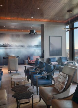 Modern waterfront lounge with plush chairs and tables, large windows over the water, and a cityscape mural on a smoky wall.