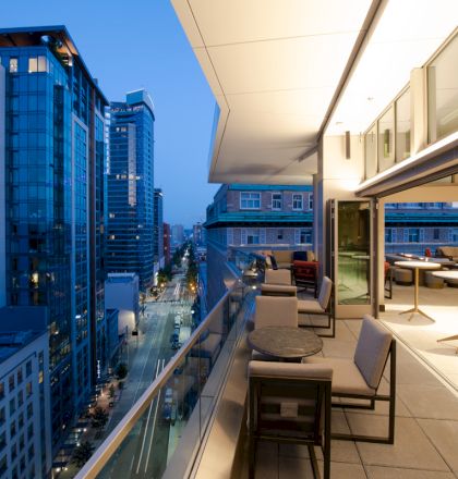 Modern high-rise city view from a rooftop terrace with lounge seating and tables at dusk.