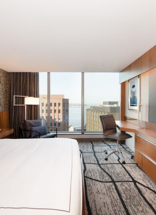 A modern hotel room with a large bed, desk chair, wall-mounted TVs, and floor-to-ceiling windows overlooking buildings.