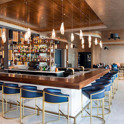 A modern hotel bar with a backlit liquor shelf, wooden counter, hanging pendant lights, and lounge seating in a bright room.