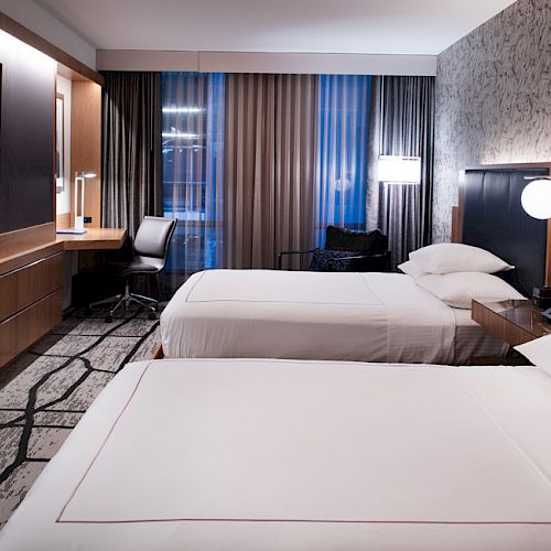 A sleek modern hotel room with two white beds, a wall-mounted TV, and blue curtains by large windows.
