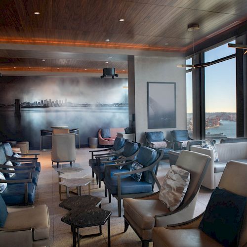 A modern luxury lounge with curved chairs and a large window overlooking a harbor and skyline, lit by warm ceiling lights.