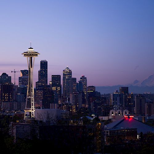 Seattle skyline at dusk with the Space Needle glowing, city lights below, and mountains silhouetted under a purple sky.