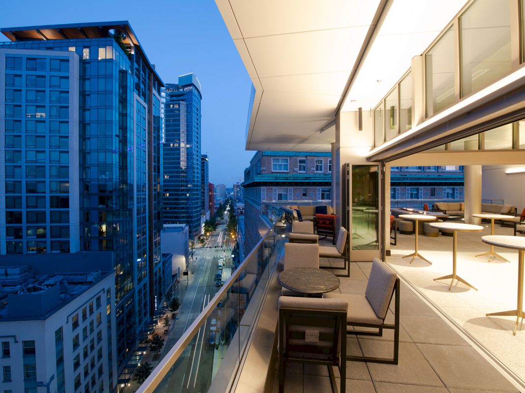 A modern high-rise terrace overlooks a city street, with lounge seating and bright sky on a clear evening.