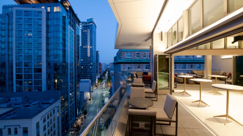 A modern high-rise terrace overlooks a city street, with lounge seating and bright sky on a clear evening.
