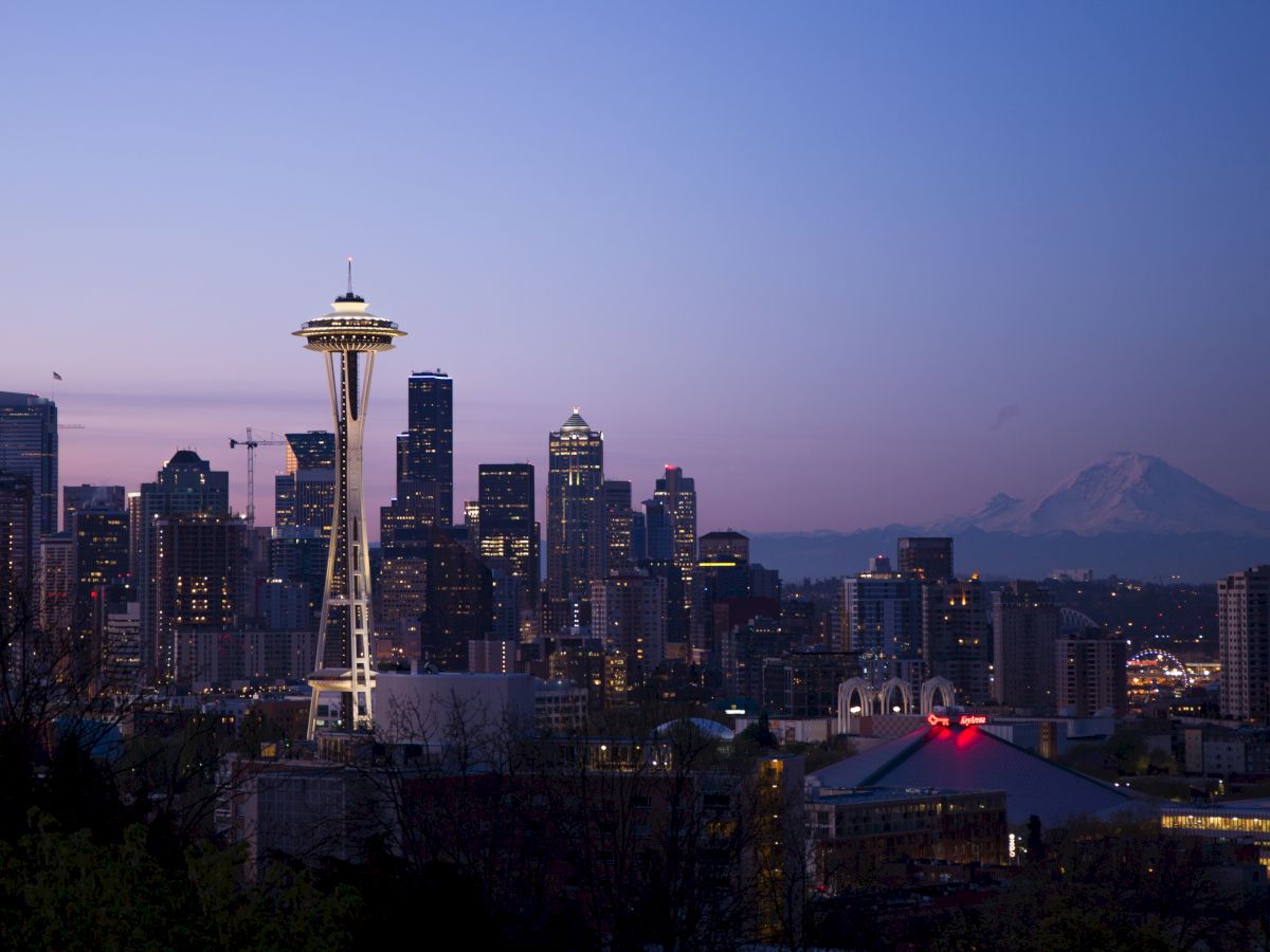 Seattle skyline at dusk: the Space Needle towers above lit buildings with mountains in the distance behind a purple sky.