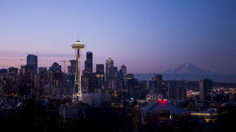 Seattle skyline at dusk: the Space Needle towers above lit buildings with mountains in the distance behind a purple sky.