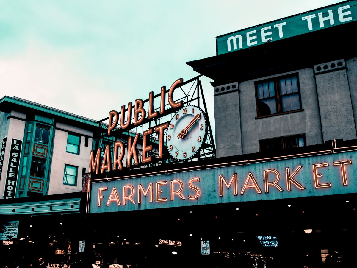 A streetfront with neon &ldquo;Public Market&rdquo; signs, a clock face, and &ldquo;FARMERS MARKET&rdquo; lettering under a blue sky.