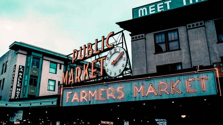 A streetfront with neon &ldquo;Public Market&rdquo; signs, a clock face, and &ldquo;FARMERS MARKET&rdquo; lettering under a blue sky.