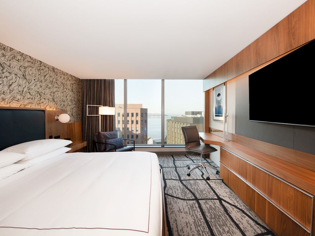 A modern hotel room with a bed, desk chair, two wall-mounted TVs, wood paneling, and large windows overlooking city buildings.