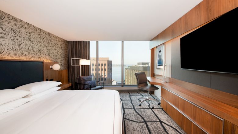 A modern hotel room with a bed, desk chair, two wall-mounted TVs, wood paneling, and large windows overlooking city buildings.