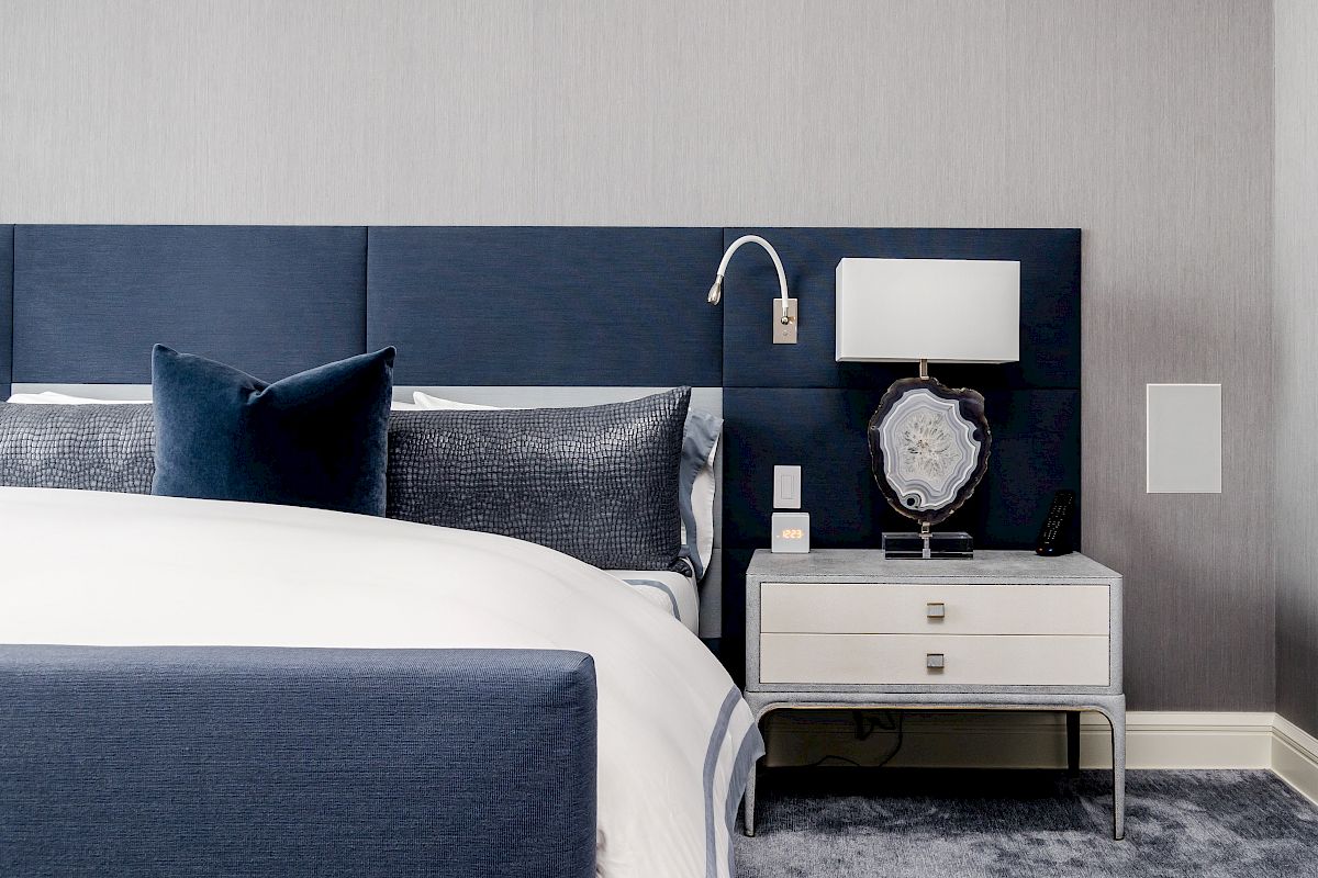 A modern hotel bedroom with a blue headboard, pillows, and a bedside nightstand topped by a lamp, clock, and decorative plate.