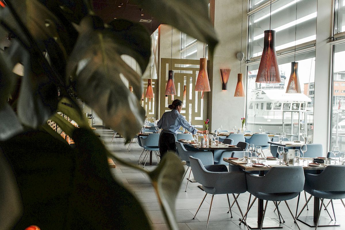 Modern restaurant dining room with blue chairs, candles and glowing pendant lamps, where a waiter sets tables by large windows.