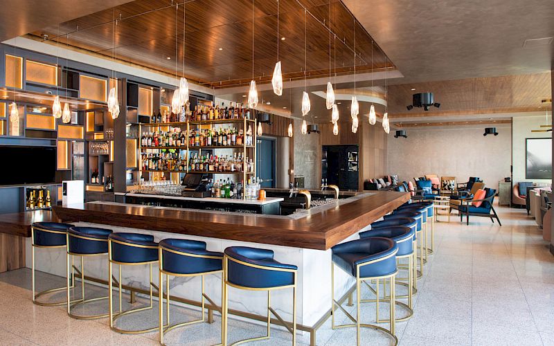 A modern bar interior with a long wooden counter, backlit shelves of liquor, hanging pendant lights, and blue stools facing a lounge area.