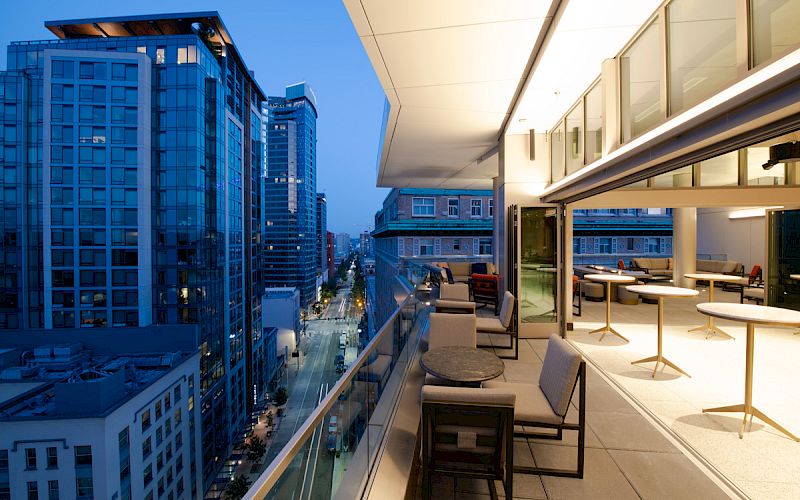 Modern rooftop terrace with seating overlooks a city street and skyline at dusk, framed by tall office towers.