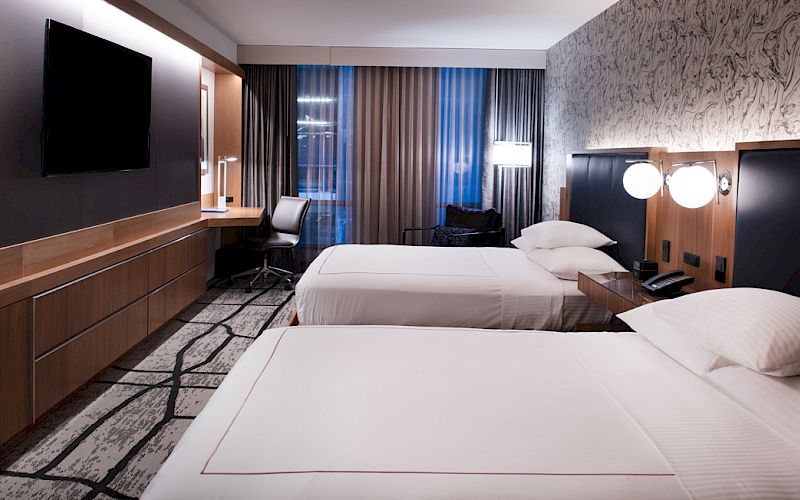A modern hotel room with two neatly made beds, a desk, curtains, and wall-mounted lights in a sleek interior.