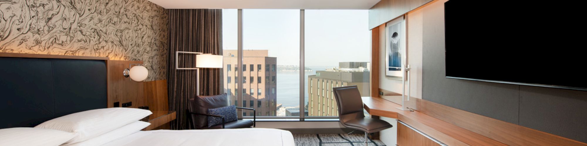 A modern hotel room with a bed, desk chair, large TVs, wood cabinetry, and city views through wide windows.