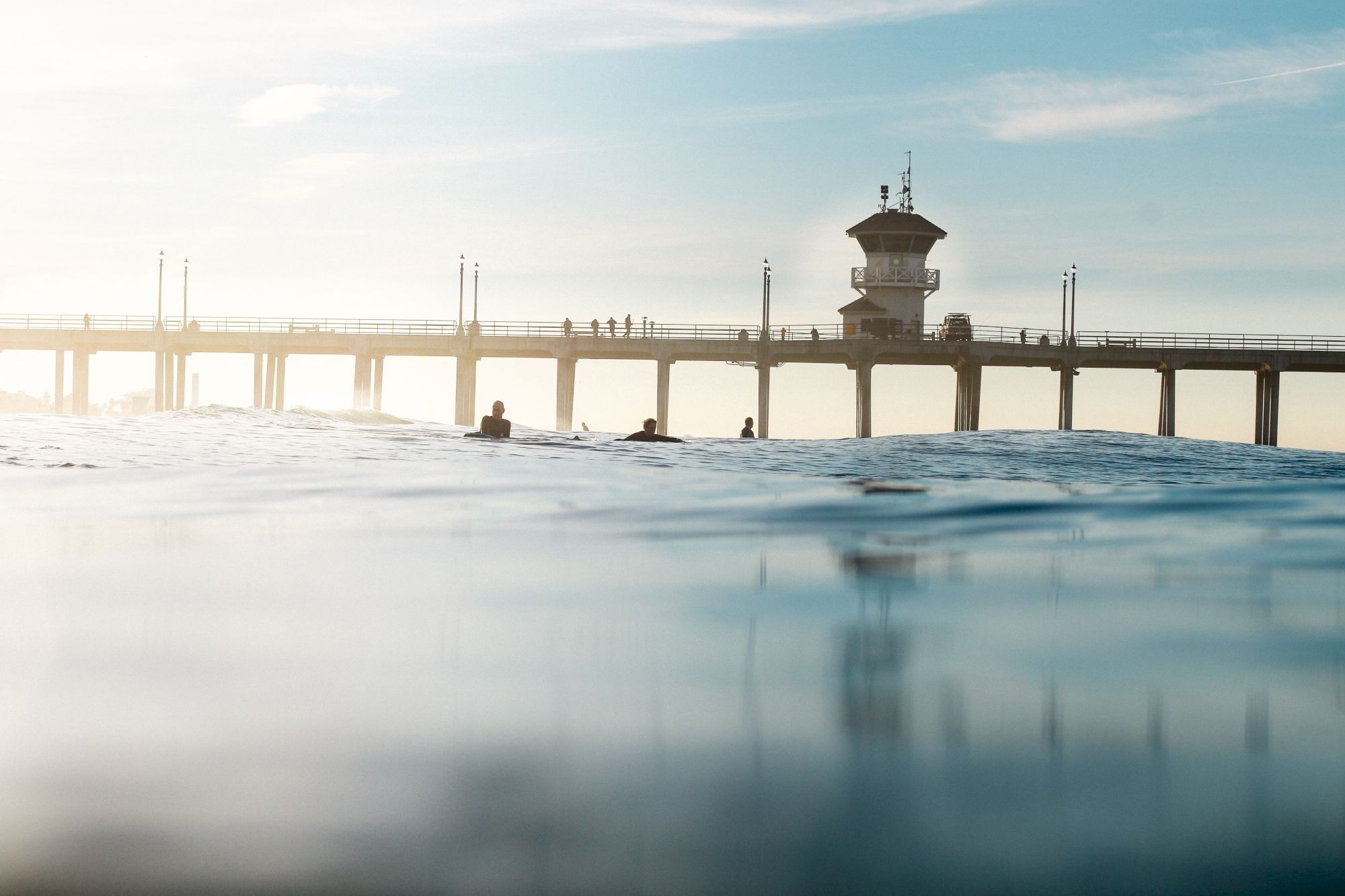 People swim in choppy water beneath a seaside pier with a lifeguard tower on a calm, cloudy day.